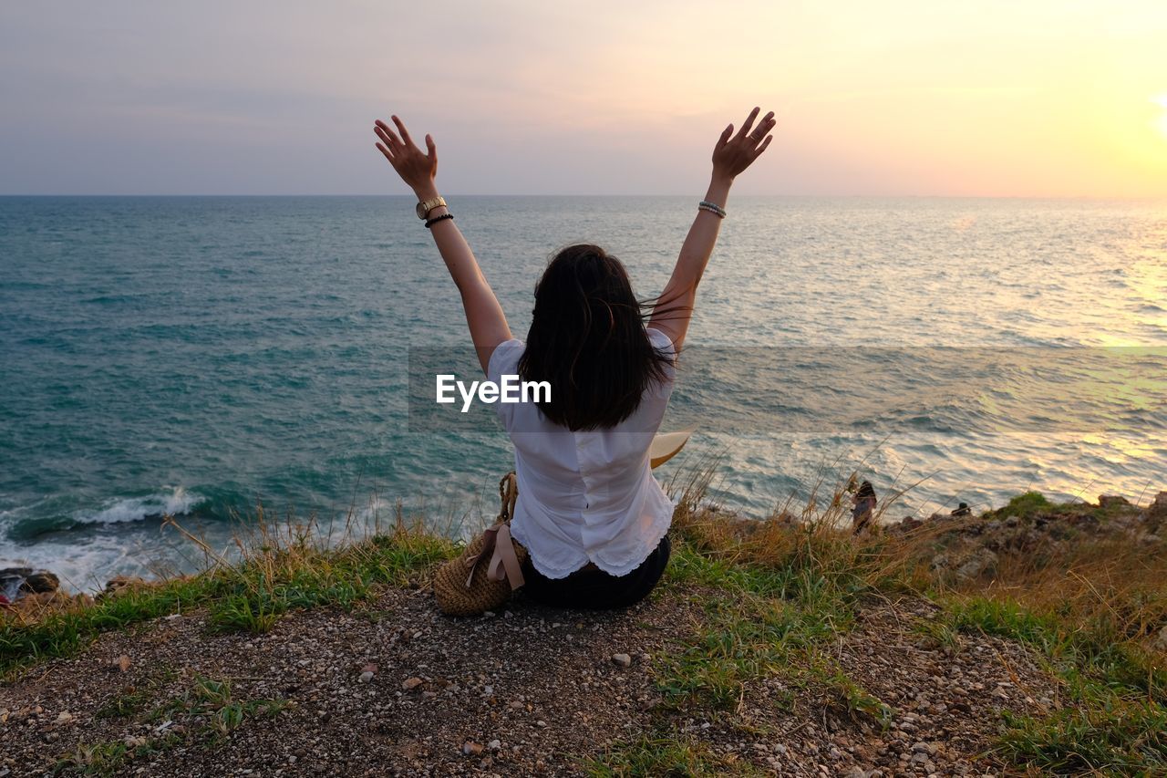 Rear view of woman with arms raised sitting at shore against sky during sunset