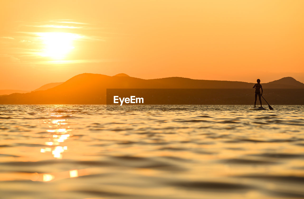 SILHOUETTE PERSON STANDING ON SEA AGAINST ORANGE SKY