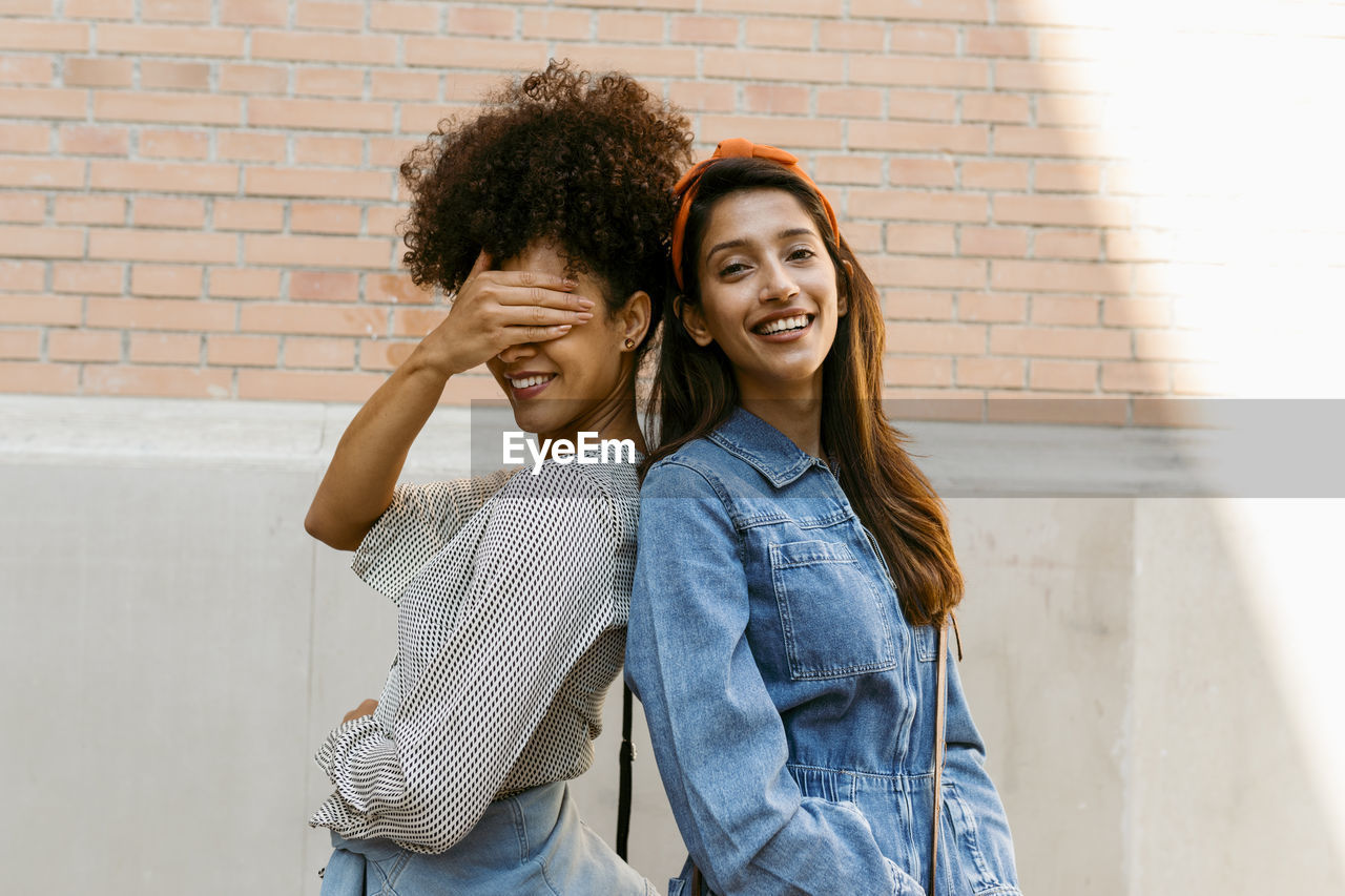 Friend covering eye with hand while standing back to back with woman against wall