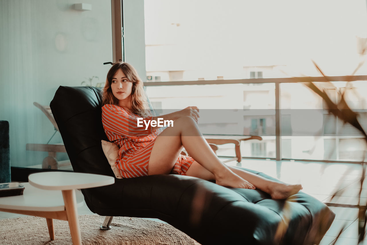 Thoughtful young woman looking away while sitting on chair against window at home