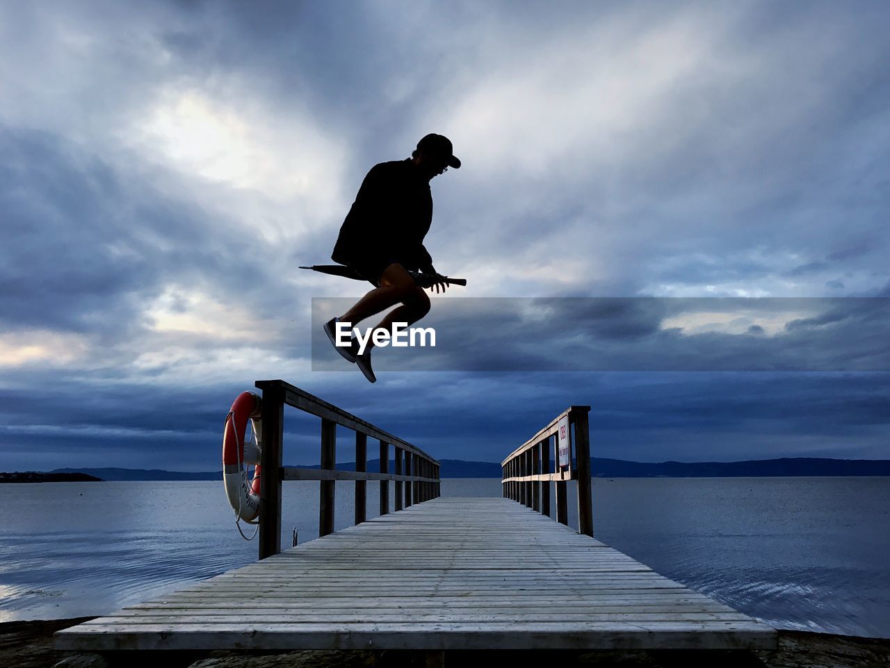 Man jumping over jetty against cloudy sky