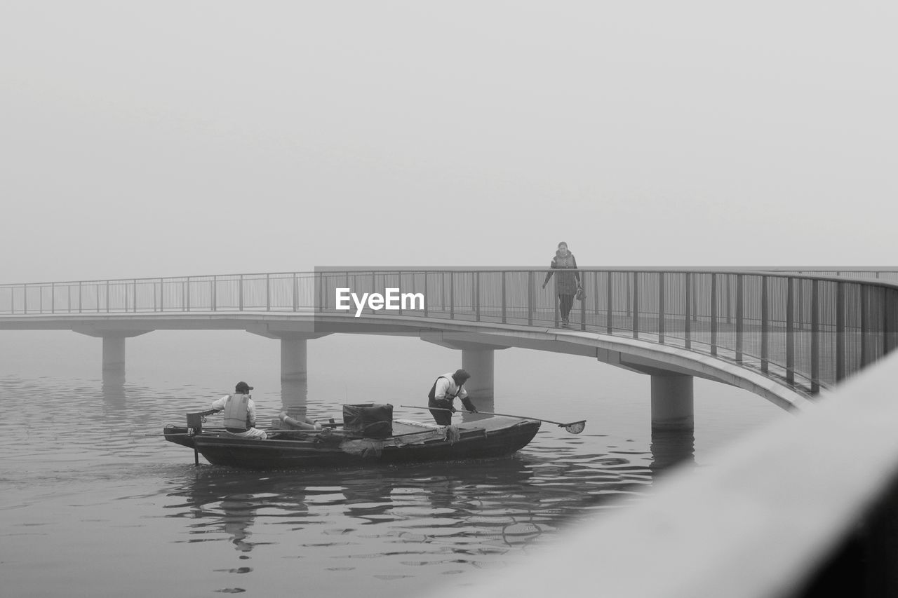 People fishing in boat on lake while woman walking on footbridge against sky