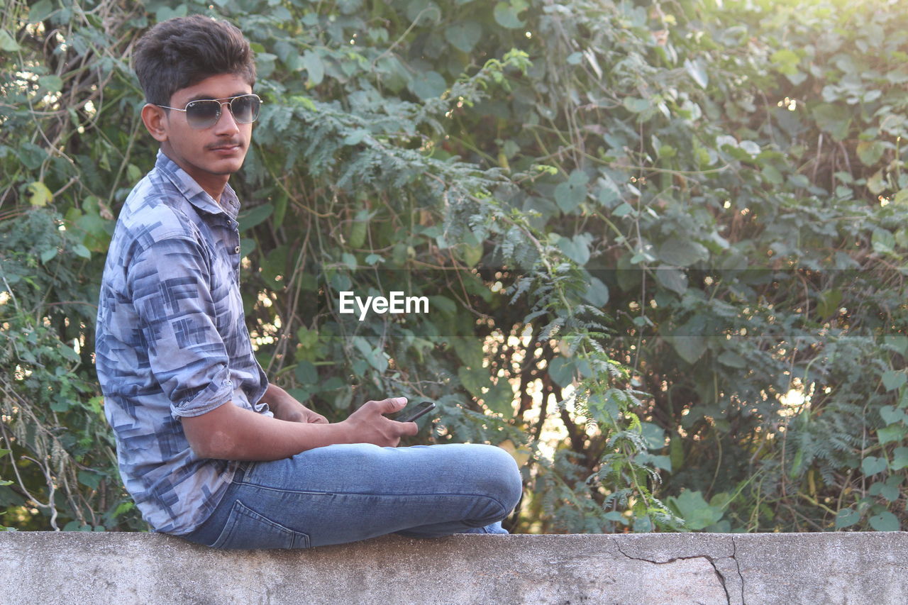 Portrait of young man wearing sunglasses while sitting against trees