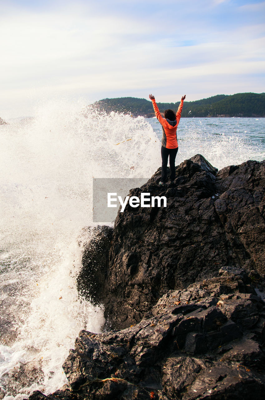 Sea waves splashing on woman standing at rocky coastline