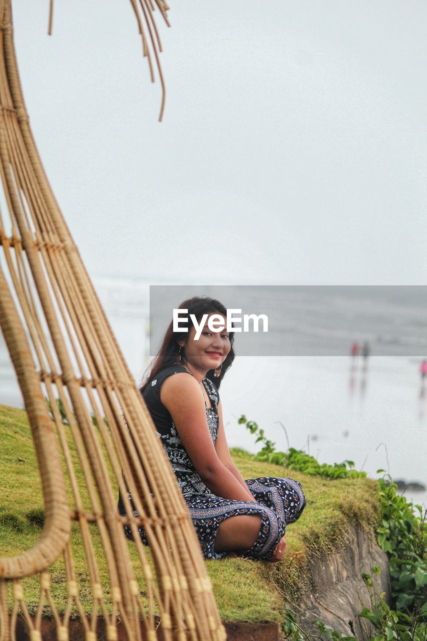 Portrait of smiling young woman sitting on shore by sea against sky