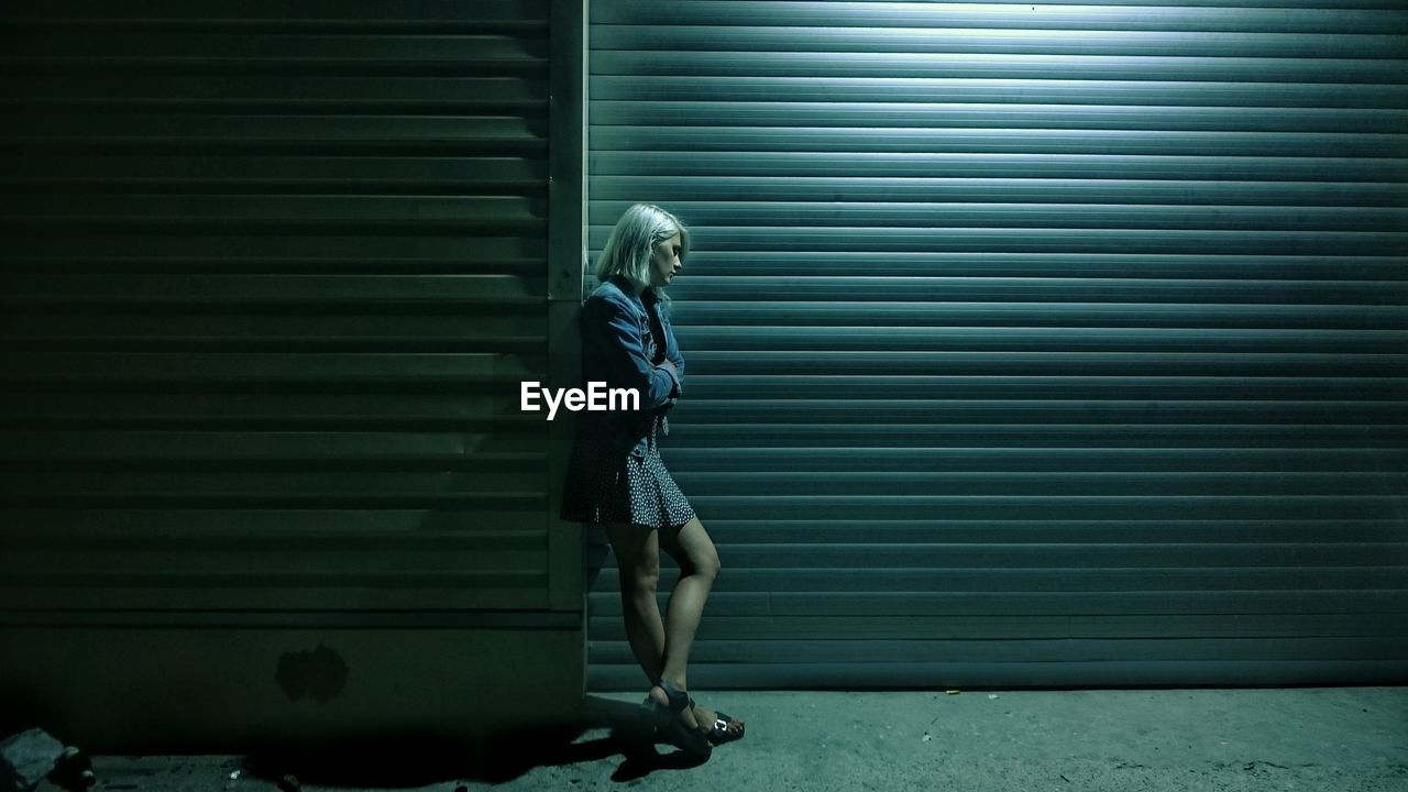 Full length of sad young woman standing by closed shutter at night