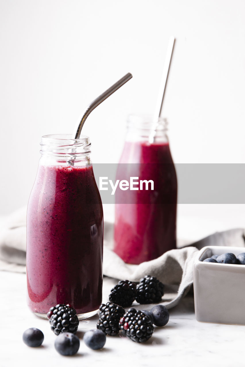 Two glass bottles of berry smoothie with metal straws, with some blackberries and blueberries on a marble surface. vertical image.