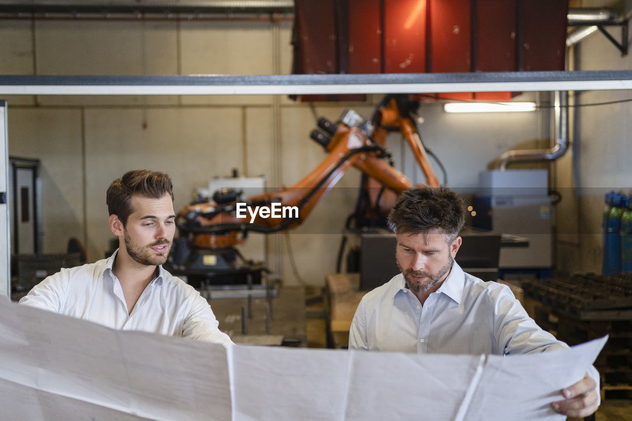 Businessmen holding blueprint paper while standing against automated machine at factory