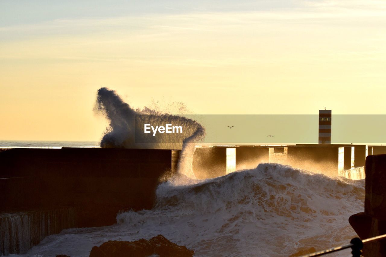 Waves splashing on shore against sky during sunset