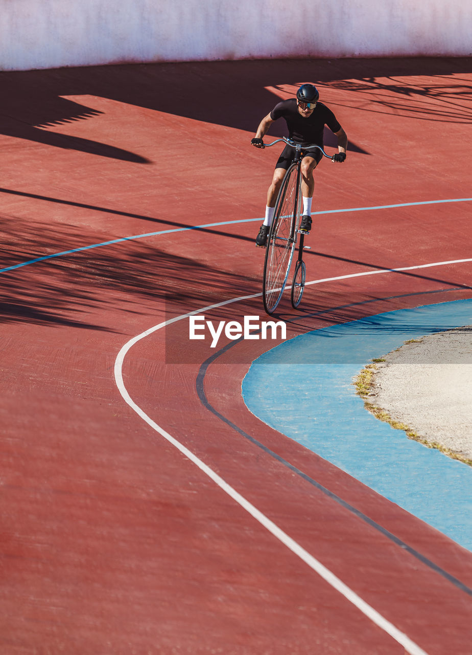 From above adult focused cyclist in black sportswear and helmet riding penny farthing along racetrack at sports stadium while getting ready for competition in sunny day