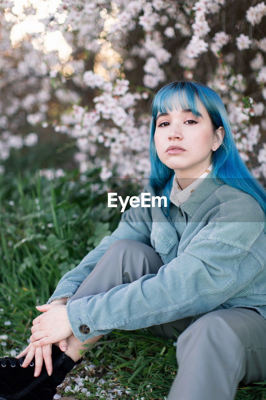 Sad millennial female model with blue hair in stylish outfit looking at camera thoughtfully while sitting on green grass near blooming tree in spring garden