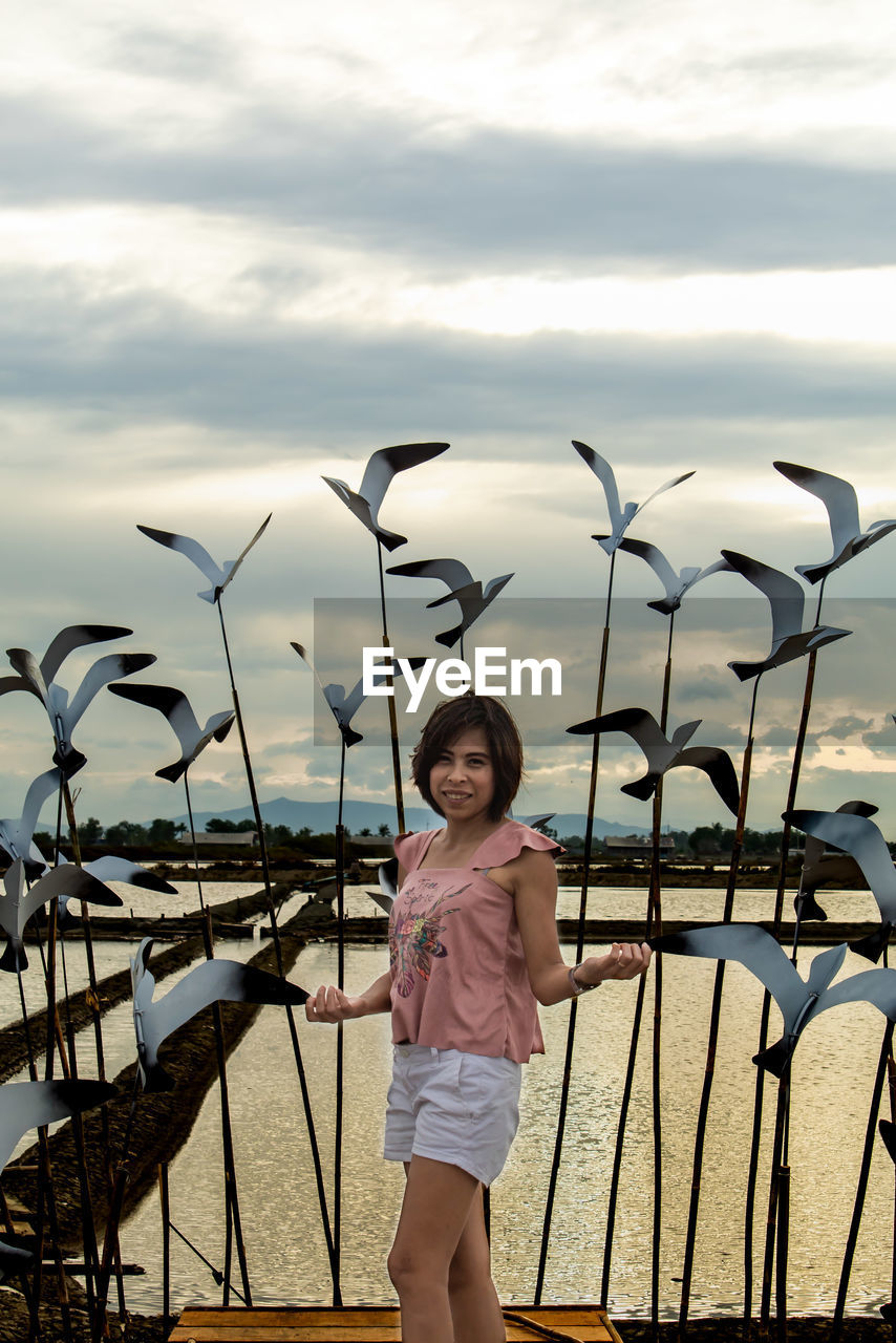 Portrait of mid adult woman standing by sculptures by lake against cloudy sky during sunset