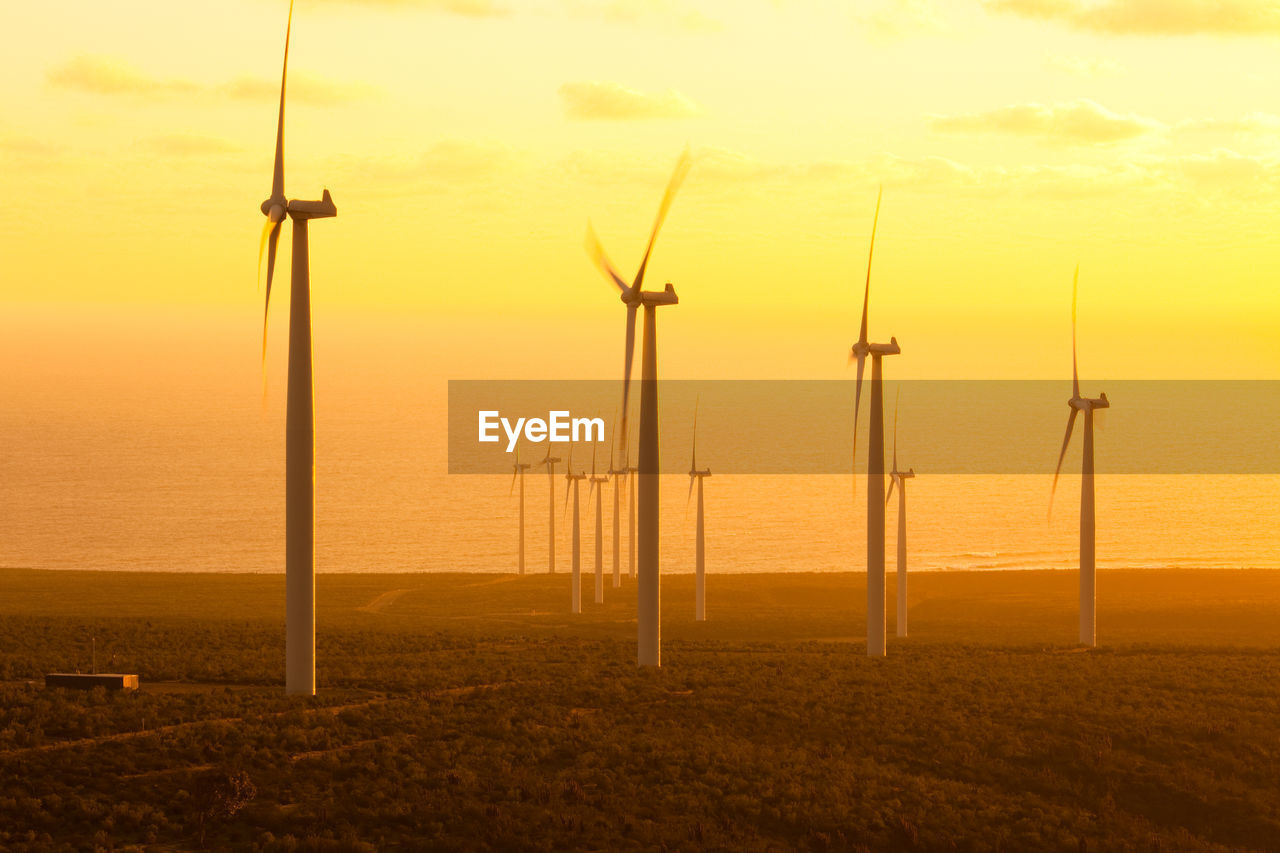 Windmills on field by sea against sky during sunset