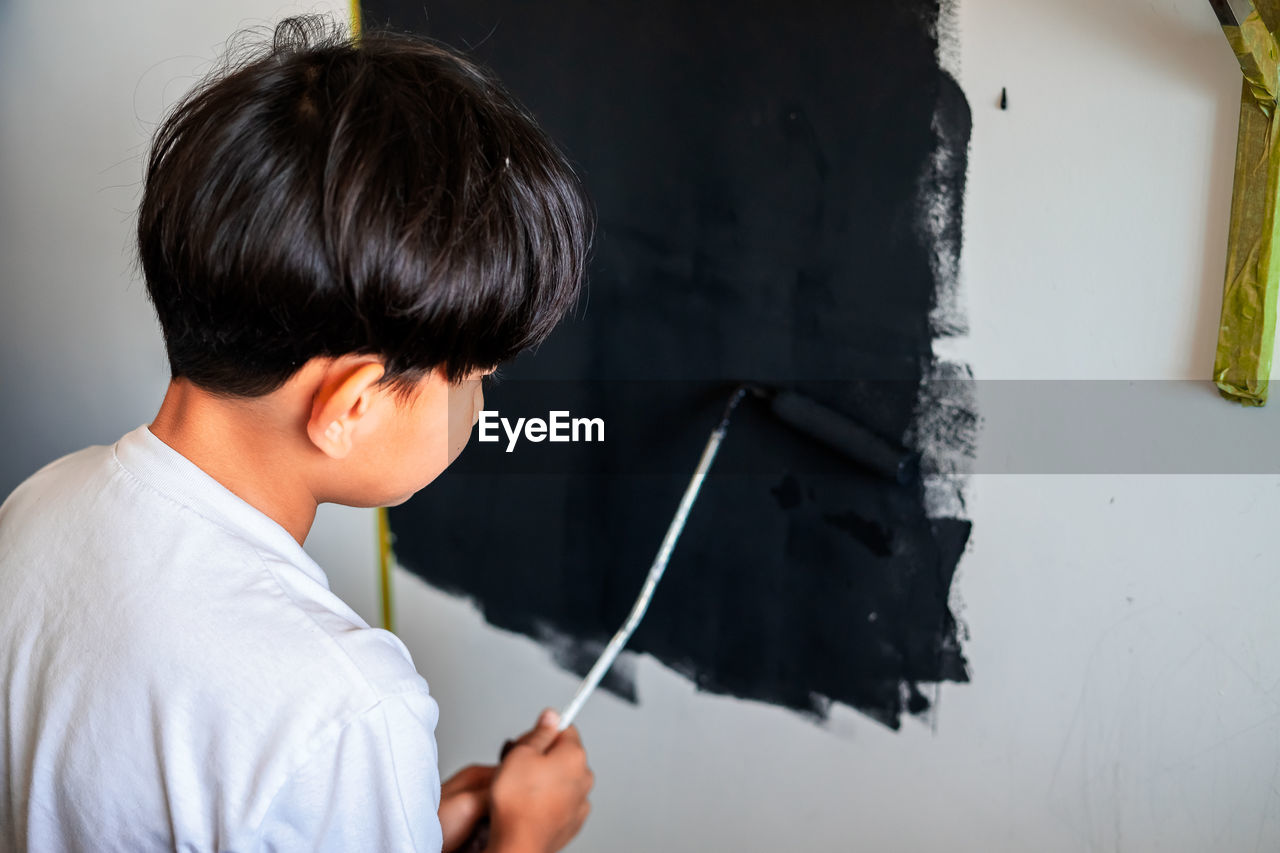 Young child painting a wall in black colour. view from behind.