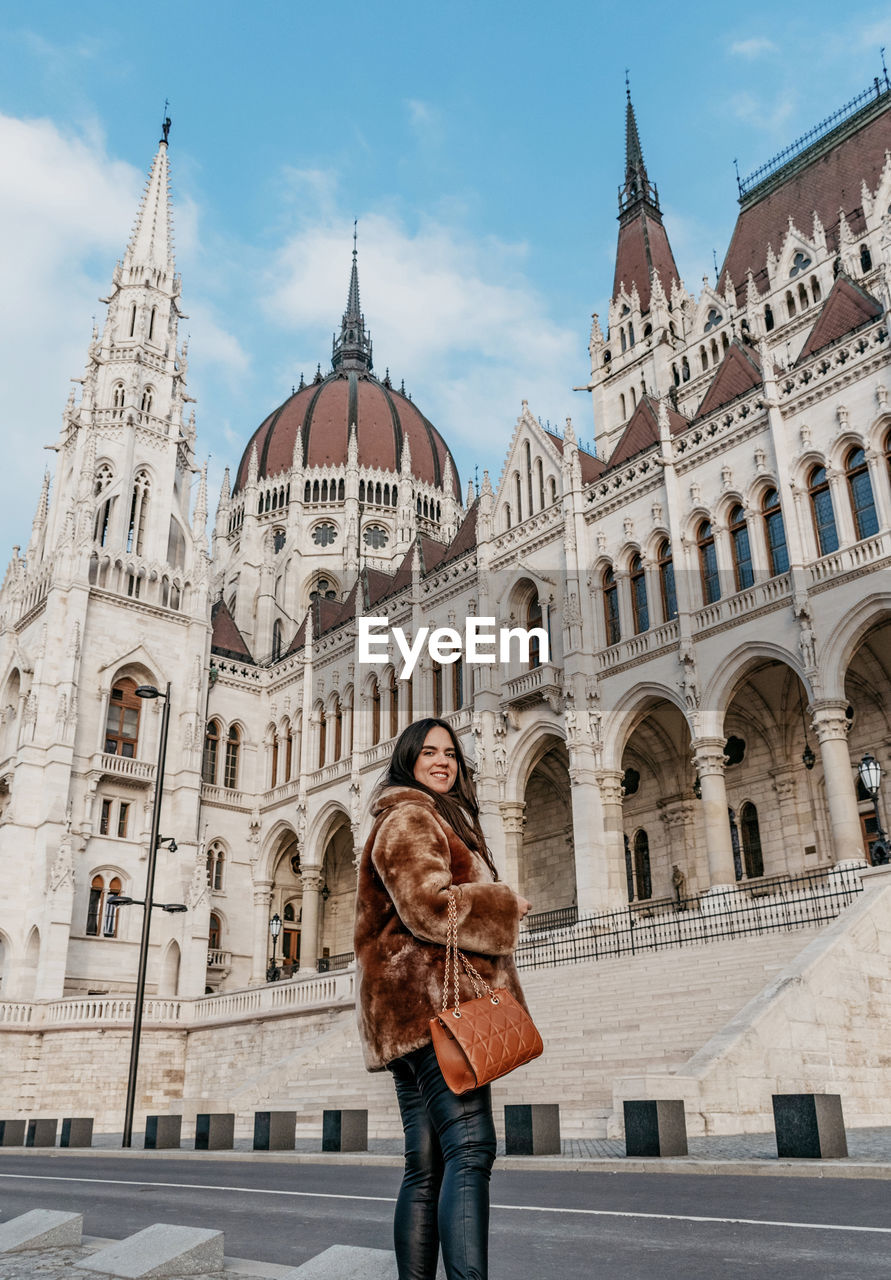 Young woman wearing stylish warm coat, standing in front of hungarian parliament in budapest hungary