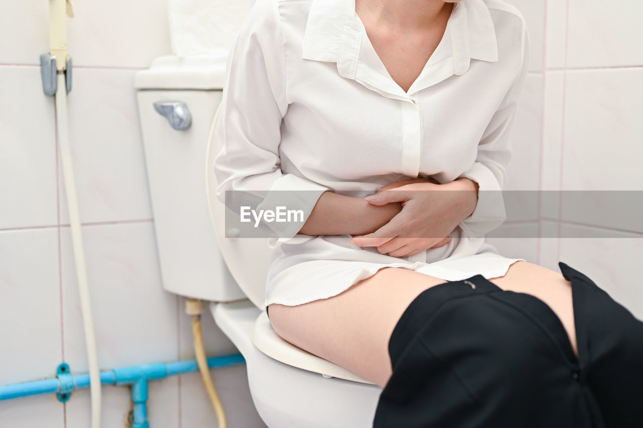 Midsection of woman sitting in bathroom while holding stomach at home