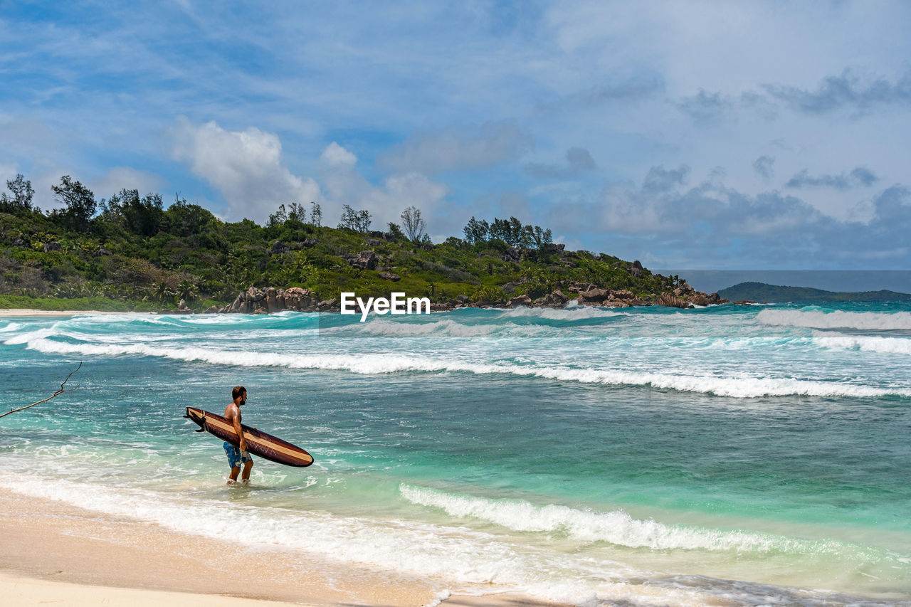 Man wearing swim trunks, standing on paradise beach, going surfing in turquoise ocean with waves