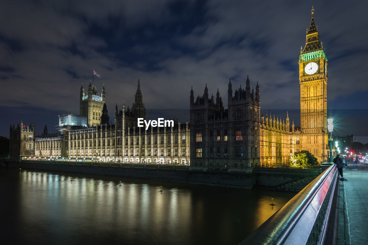 Thames river by illuminated big ben and houses of parliament against sky at night