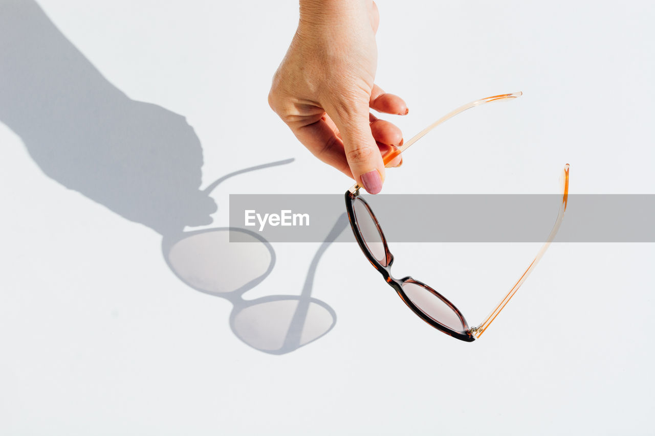 From above cropped anonymous female hands holding trendy sunglasses with brown lens and frame over white background with shadow