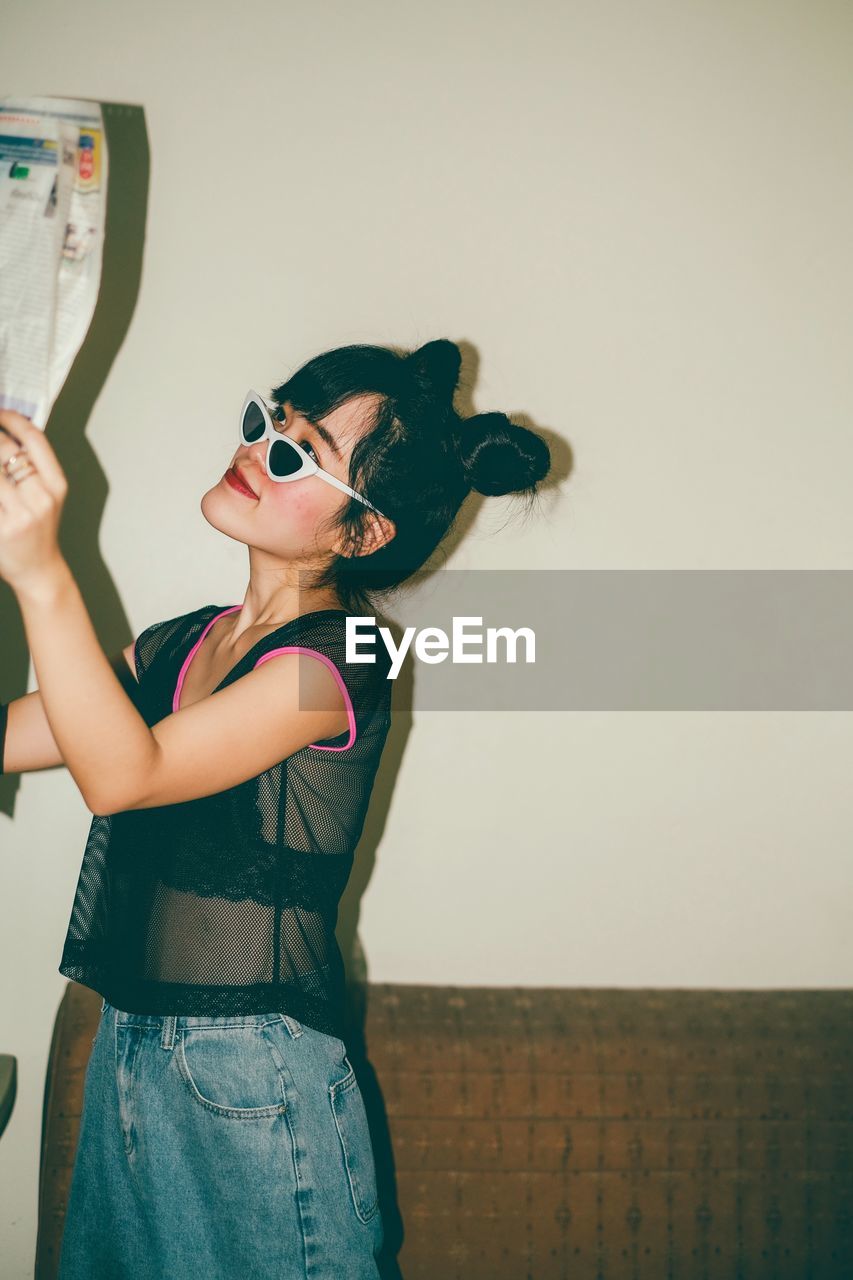 MIDSECTION OF WOMAN HOLDING SUNGLASSES STANDING AGAINST WALL