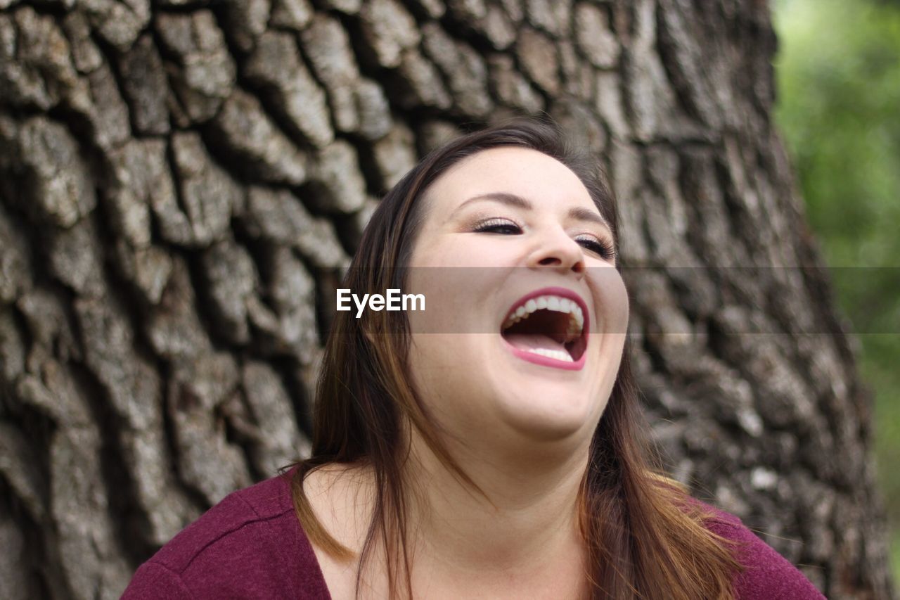 Close-up of young woman looking away while laughing against tree trunk
