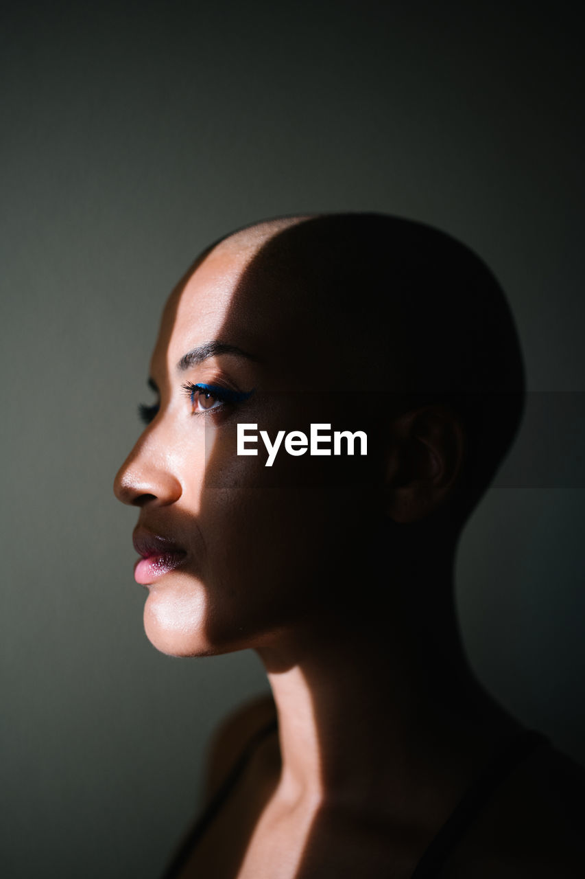 Side view of thoughtful african american female with bald hairstyle standing near window in apartment and illuminated by sunbeam in morning