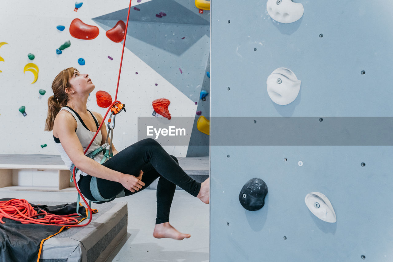 Side view of active female alpinist with belay looking up near artificial climbing wall with grips during workout in modern gym