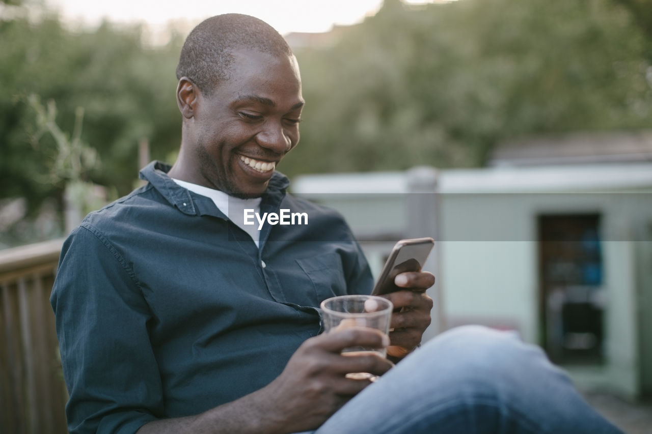 Smiling mid adult man using mobile phone while holding glass at porch