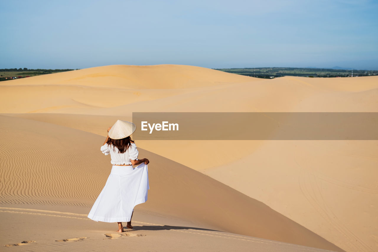 Rear view of woman wearing hat walking on sand dune at desert against sky