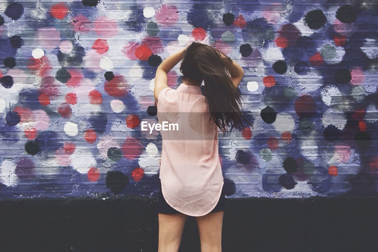 Rear view of woman with arms raised holding hair against graffiti wall