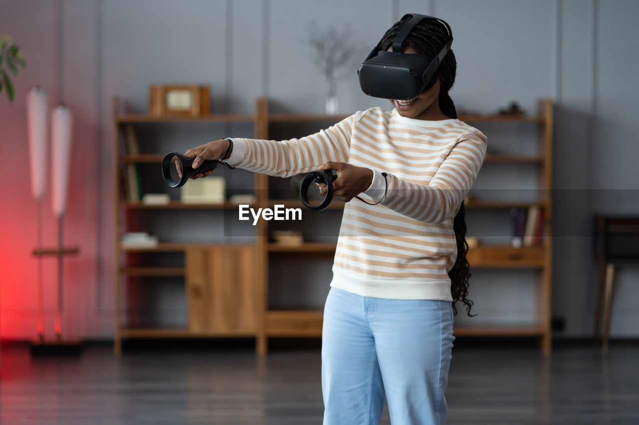 Happy young african american woman wearing virtual reality goggles playing video games at home