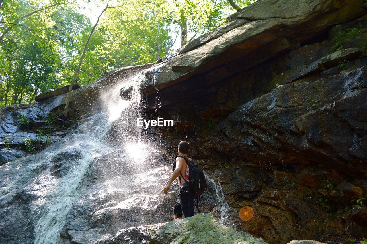 Low angle view of shirtless man standing on rock by waterfall