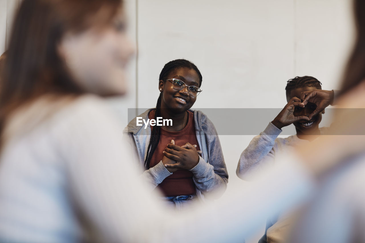 Smiling teenage girl sitting with hands on chest by male friend gesturing heart shape and looking at students in group t