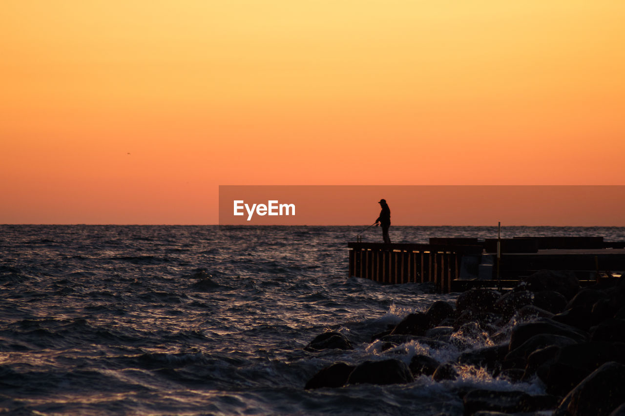 Silhouette man fishing on pier over sea against orange sky