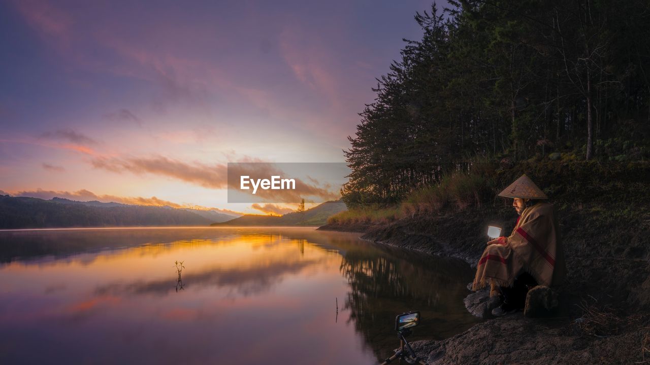 Man in hat sitting at lakeshore against sky during sunset