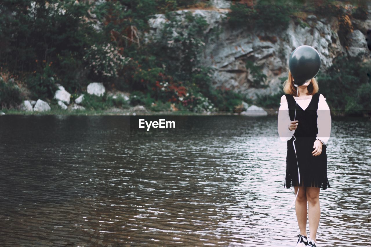 Full length of woman standing by lake and holding black balloon