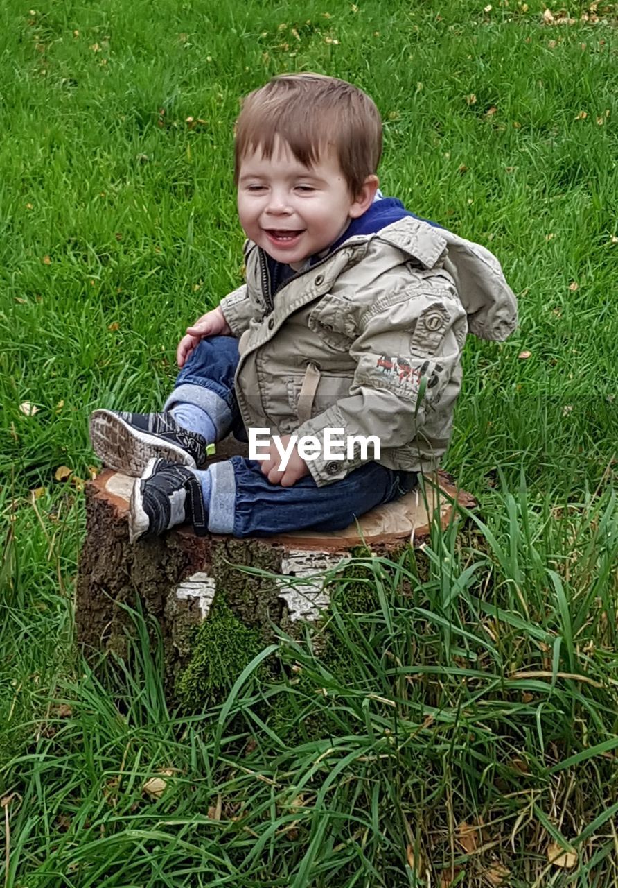 Cheerful baby boy sitting on tree stump amidst grassy field