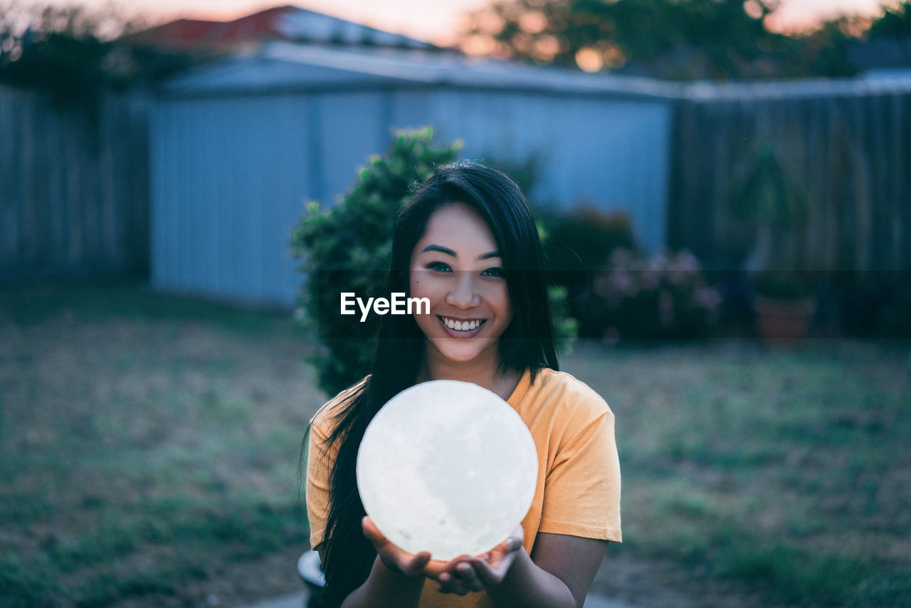 Portrait of smiling young woman holding illuminated ball on field during sunset