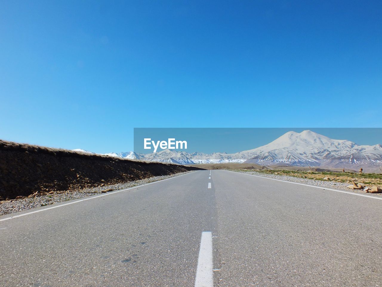 road, sky, mountain, transportation, scenics - nature, the way forward, landscape, snow, environment, nature, blue, beauty in nature, cold temperature, road marking, no people, winter, clear sky, asphalt, marking, diminishing perspective, highway, day, symbol, horizon, travel, tranquil scene, empty road, non-urban scene, mountain range, land, tranquility, travel destinations, copy space, vanishing point, sign, remote, snowcapped mountain, sunny, outdoors, road trip, idyllic, country road, infrastructure, road surface, journey