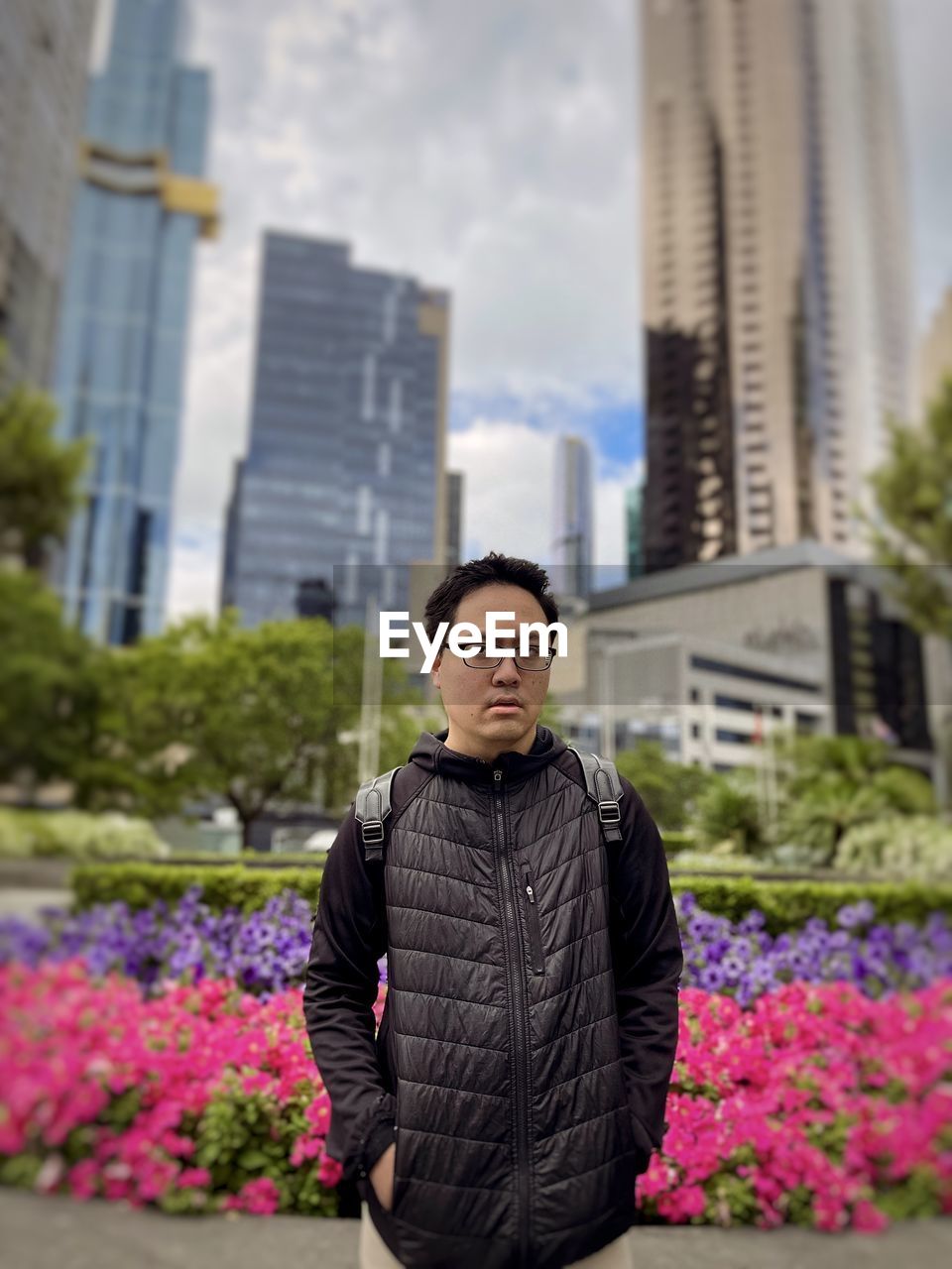 Asian man standing against flower beds, trees and skyscrapers in the city.