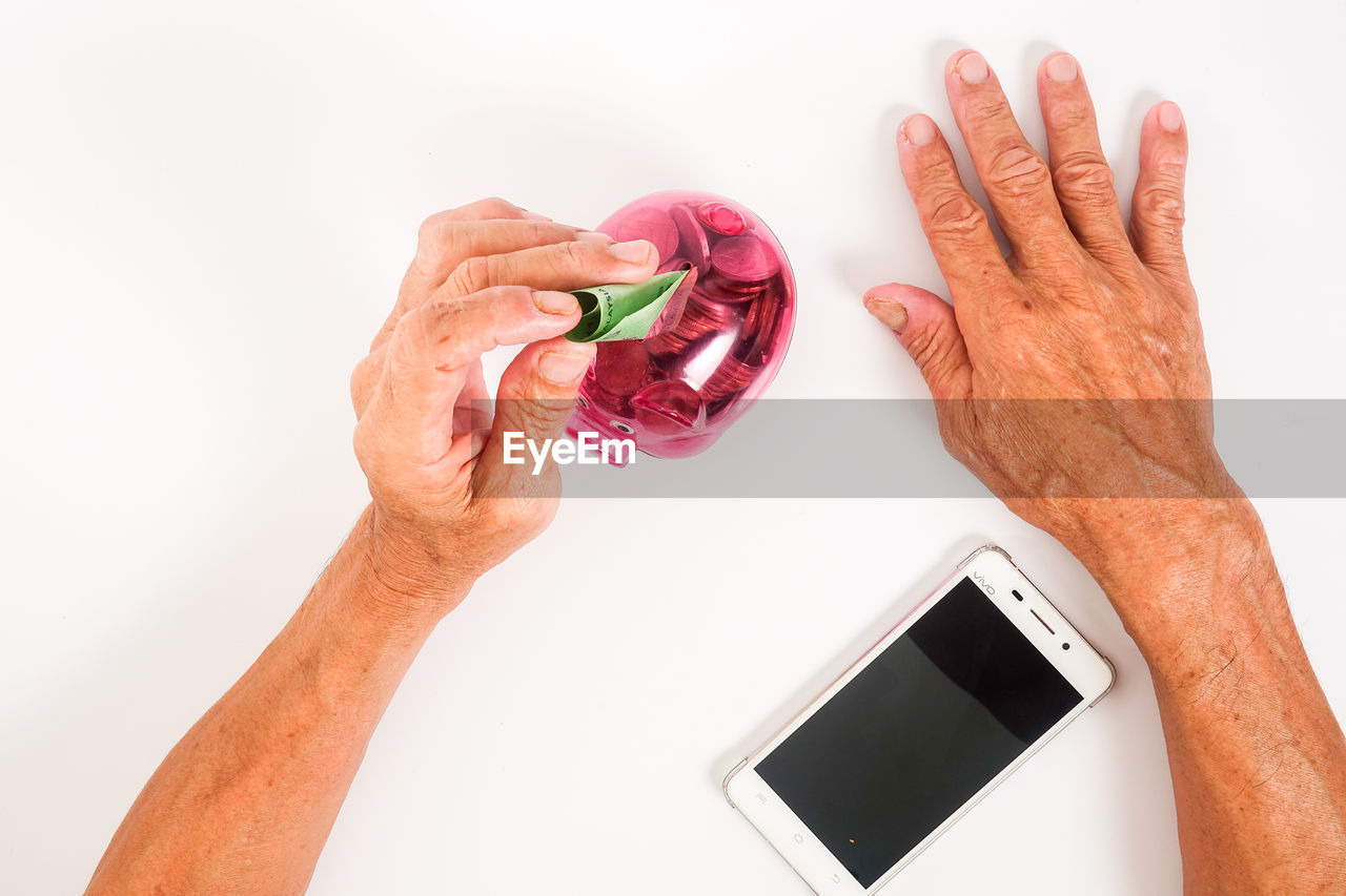 Close-up of man inserting money in piggy bank with mobile phone against white background