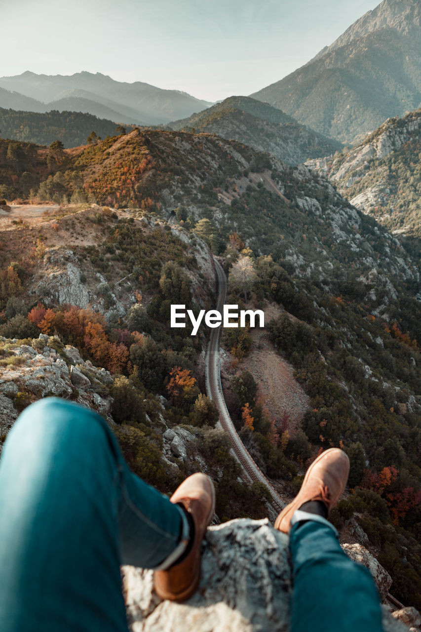 Low section of man sitting on cliff against mountains