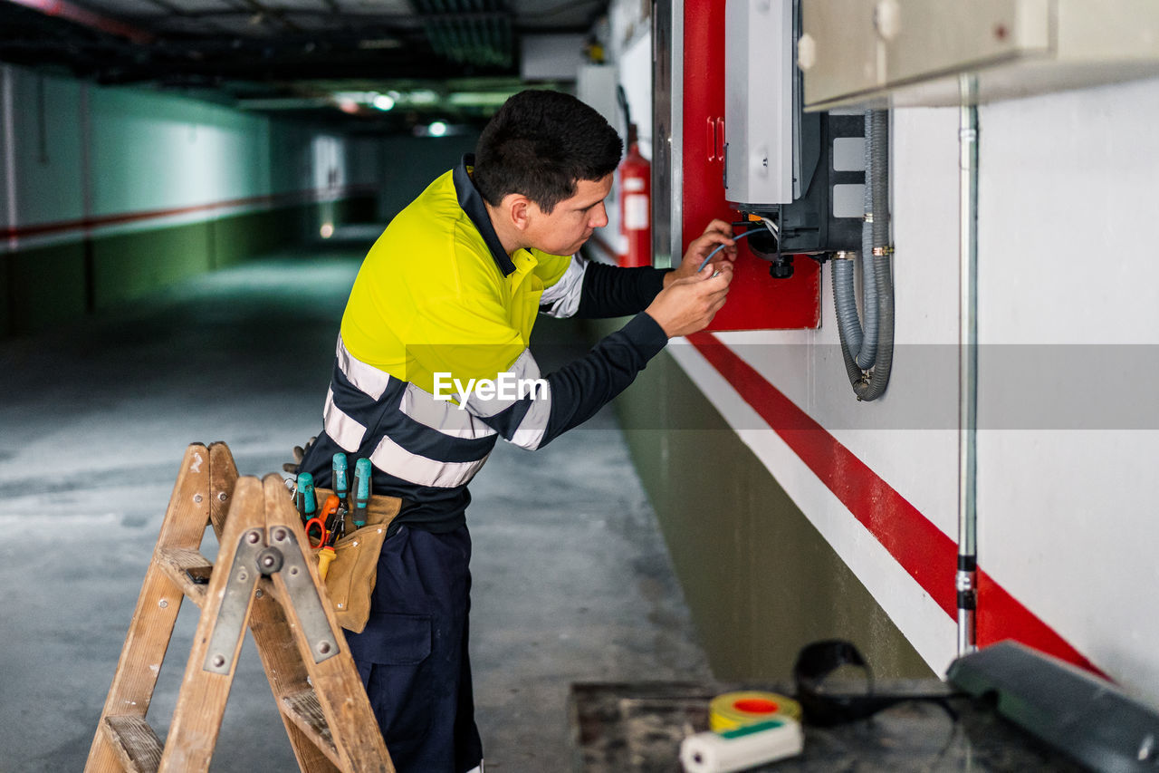 Side view of young professional male technician with electric tools repairing and checking equipment while working in building