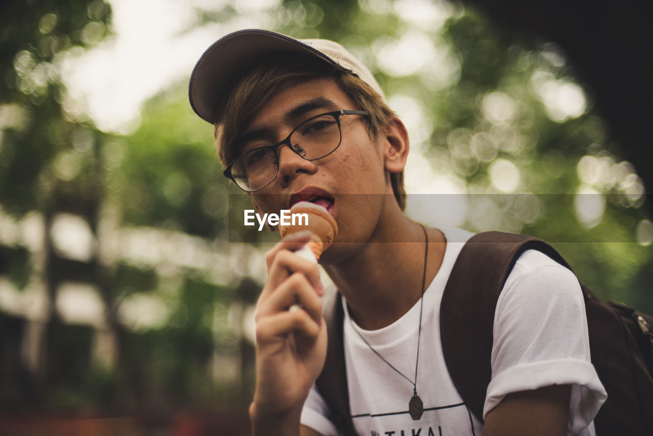 Portrait of young man eating ice cream cone while standing against trees