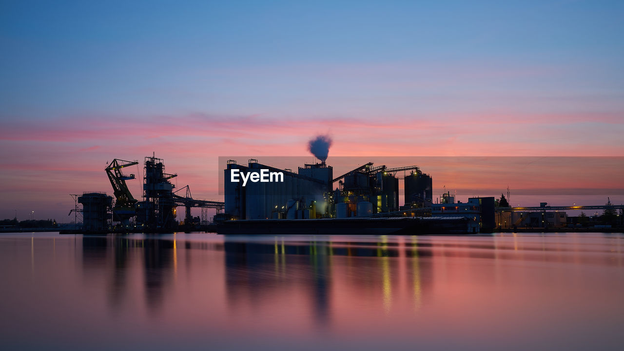 Industrial buildings by river against sky during sunset