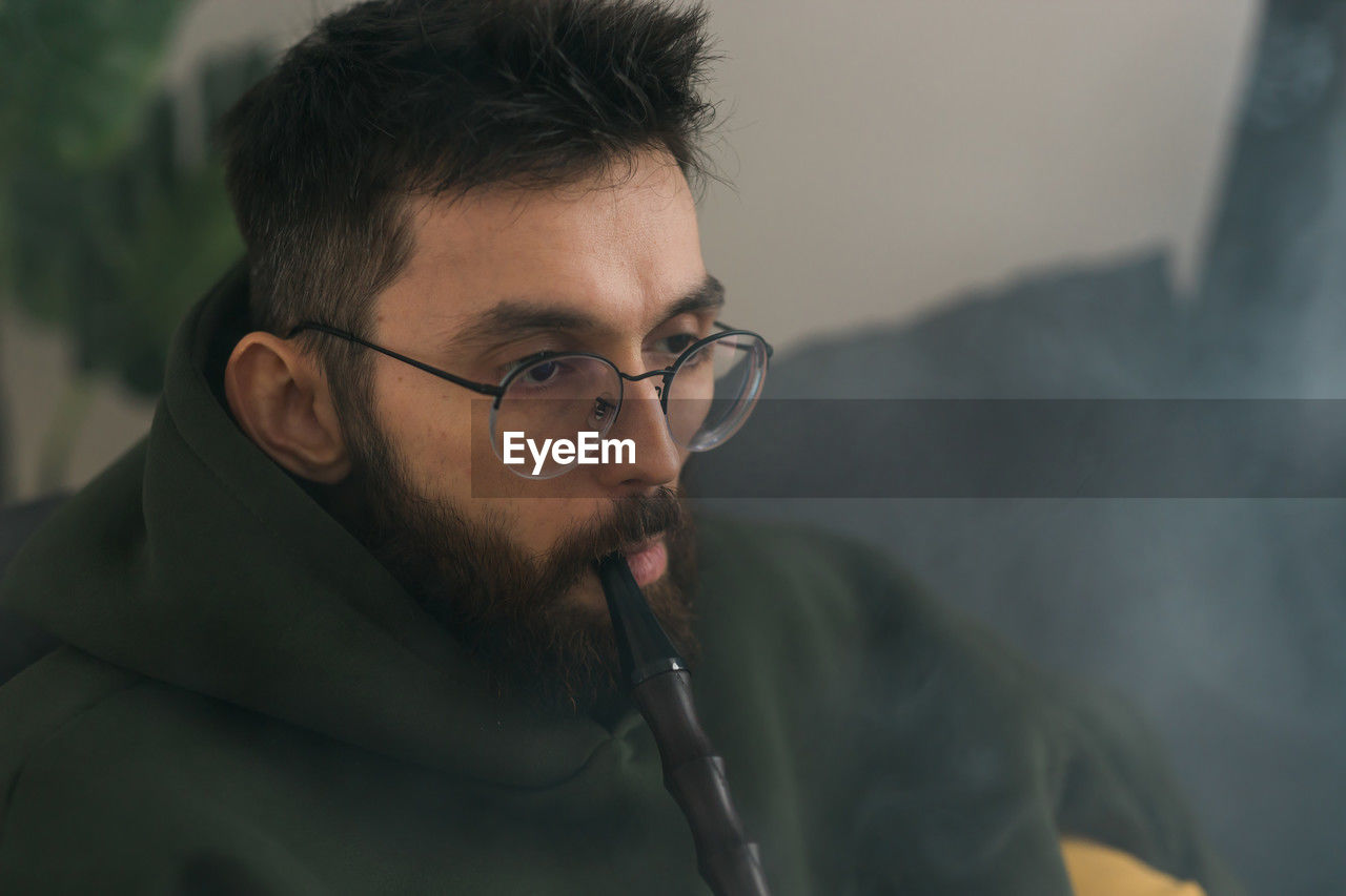eyeglasses, one person, glasses, adult, men, beard, portrait, facial hair, headshot, person, indoors, looking, smoke, black hair, lifestyles, close-up, young adult, mature adult, activity