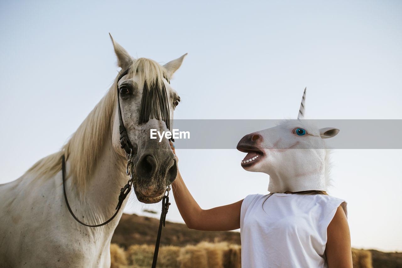 Young woman wearing unicorn mask gently stroking white horse