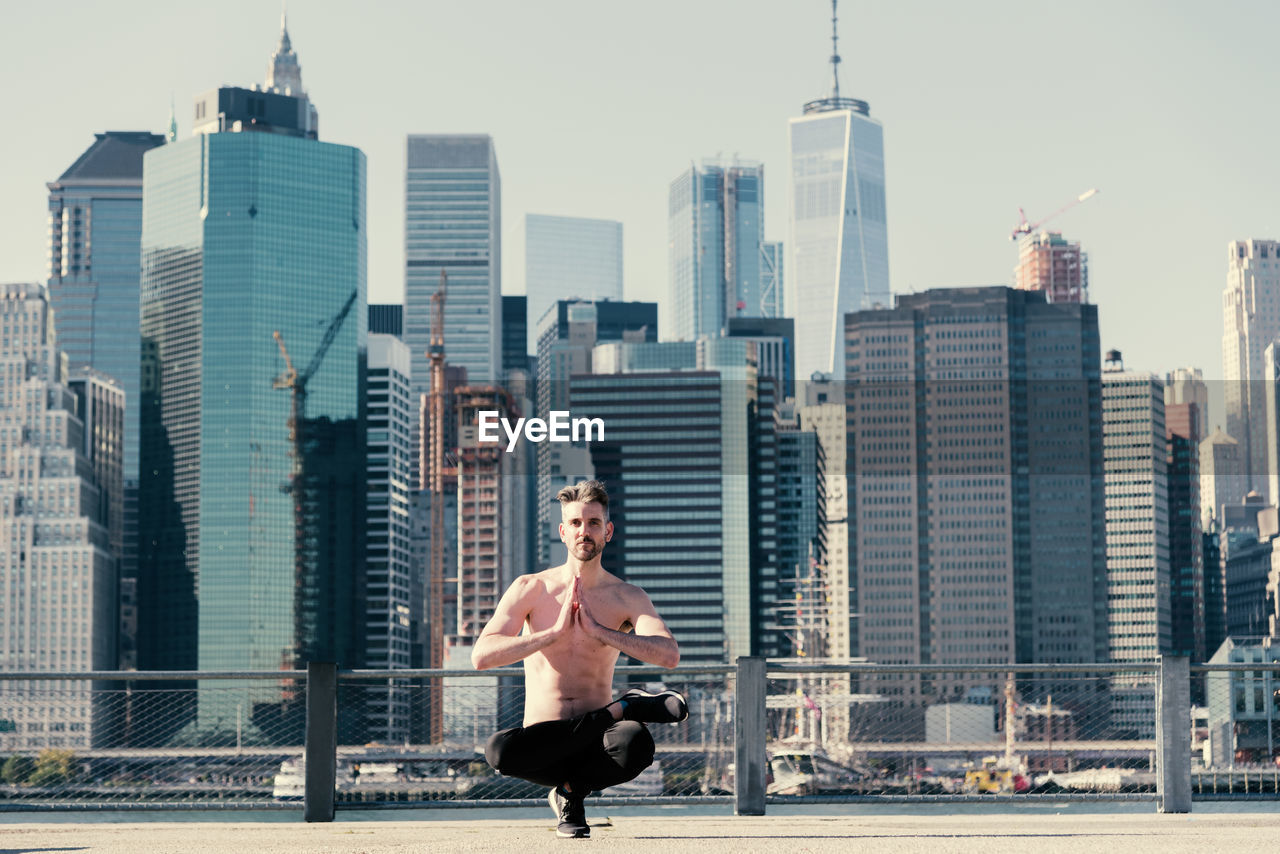 Full length of shirtless mature man practicing yoga on building terrace against one world trade center
