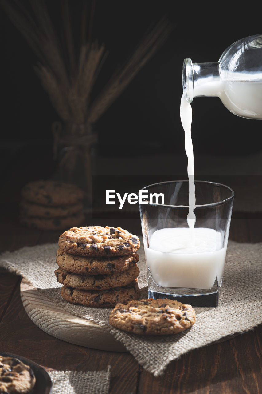 Chocolate chip cookies and milk bottle spilling milk in glass on wooden base, dark background. 