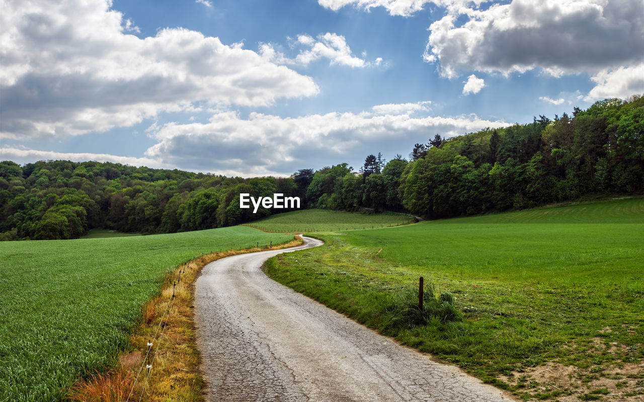 Narrow road amidst grassy field against cloudy sky